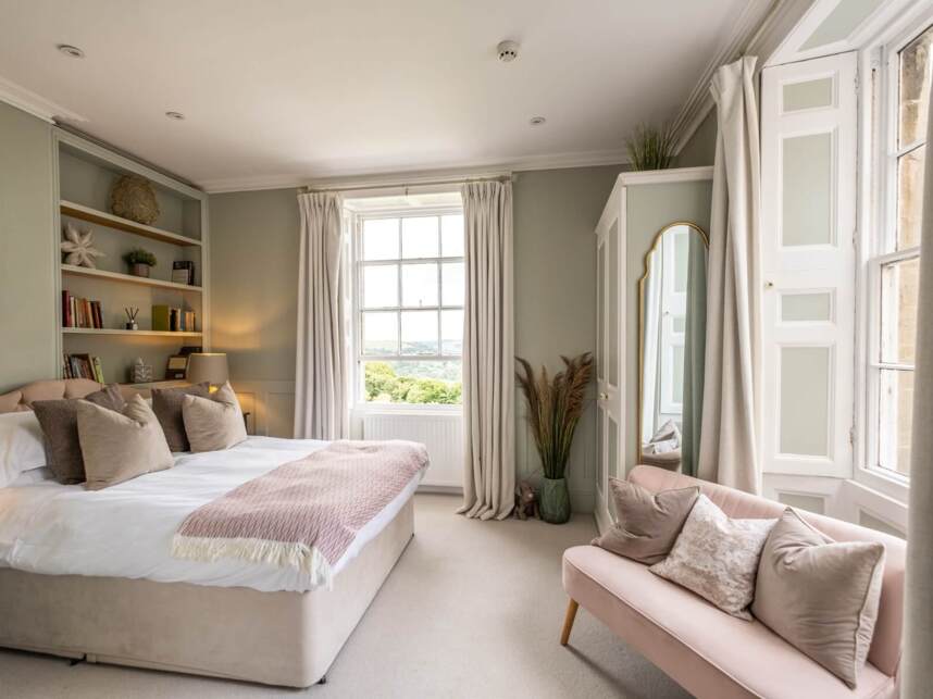 Bright hotel bedroom with large window, soft beige bed with multiple pillows, built-in shelves, pink loveseat, and elegant full-length mirror