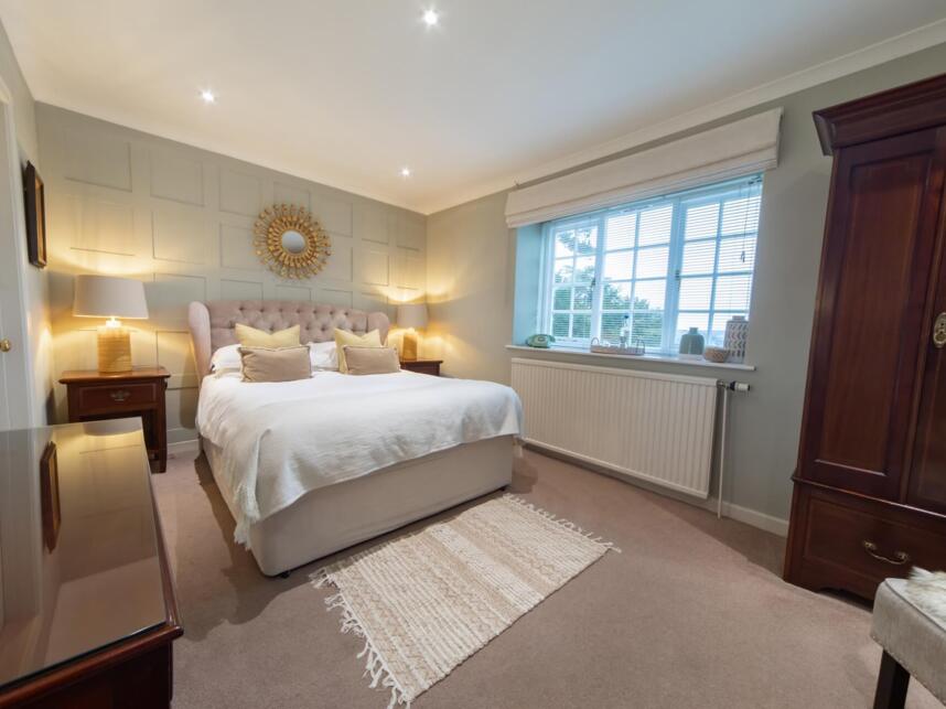 Cozy bedroom with tufted king bed, soft lighting, decorative mirror, wooden wardrobes, window with blinds, and neutral decor