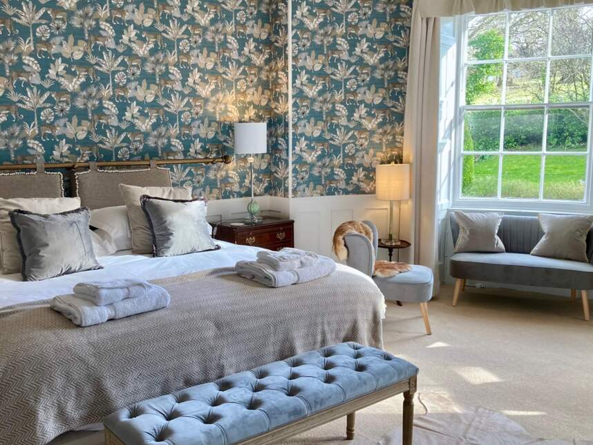 Elegant hotel bedroom with floral patterned wallpaper, king-size bed, velvet bench, cozy seating, and large window overlooking a green garden