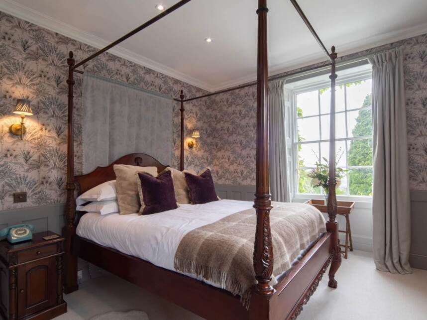Elegant vintage-style bedroom with carved wooden four-poster bed, plush pillows, floral wallpaper, large window with garden view