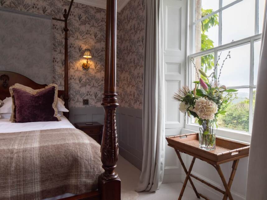 Cozy guest room with carved wooden four-poster bed, plush velvet pillows, floral wallpaper, and fresh flowers by a large window with countryside views