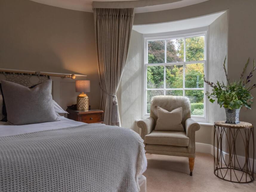 Cozy hotel room with beige armchair by large window, soft bedding, wooden nightstand with lamp, and floral arrangement on side table