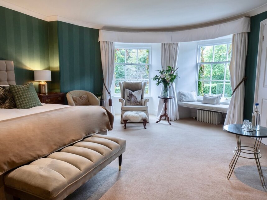 Spacious hotel bedroom with plush beige bedding, green striped walls, seating area by large windows, and elegant décor