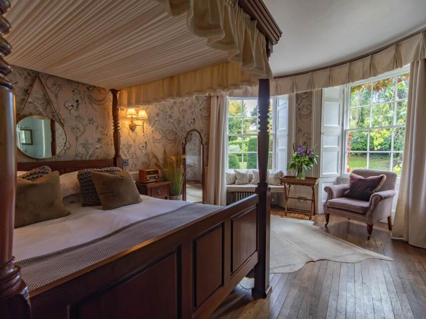 Cozy bedroom with four-poster bed, plush pillows, floral wallpaper, vintage radio, armchair, and garden views through large windows