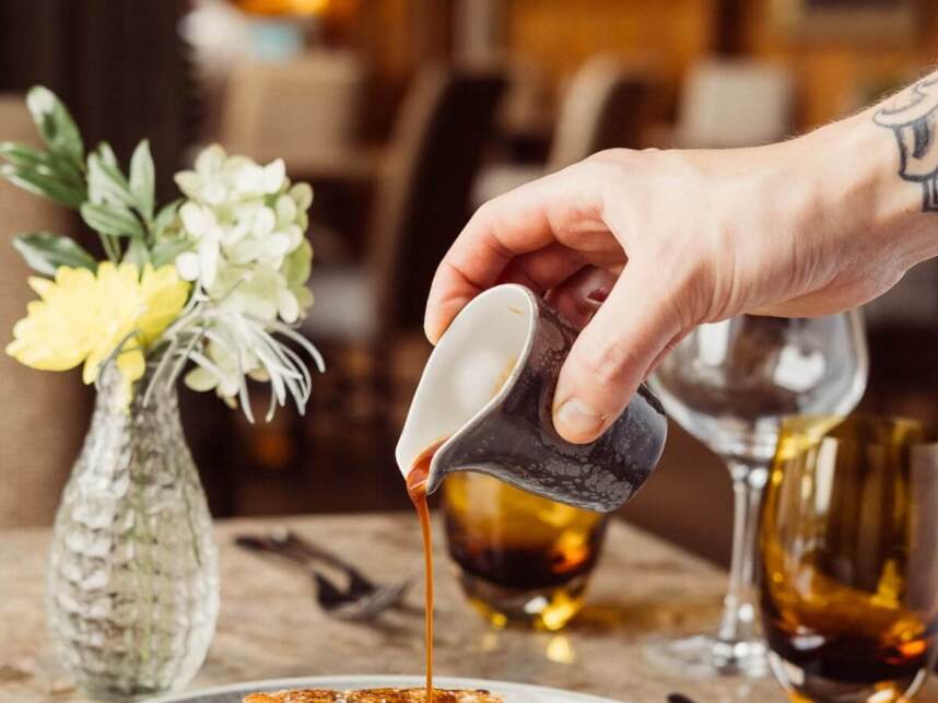 Hand pouring sauce over a plated gourmet pork dish with roasted vegetables in an elegant dining setting with glassware and fresh flowers