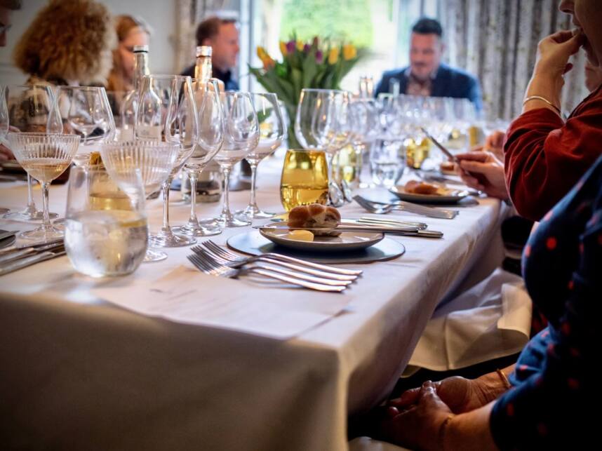 Elegant dining table set with multiple wine glasses, bread rolls, and butter, guests engaged in conversation in a cozy restaurant.