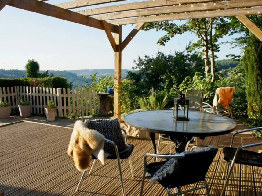 Sunny wooden terrace with round table, metal chairs draped with cozy throws, overlooking lush green hills and garden plants