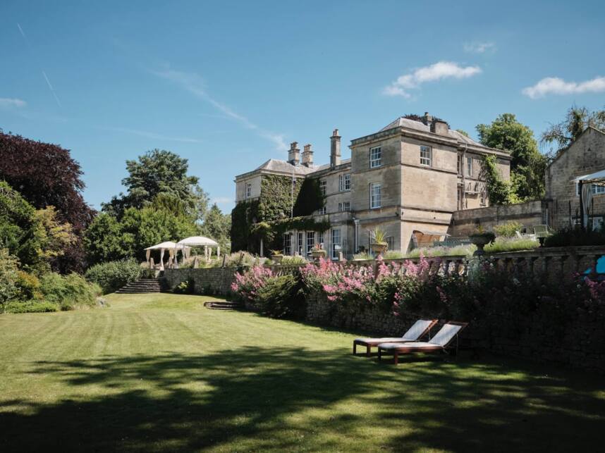 Historic stone hotel surrounded by lush gardens, pink flowers, and sun loungers on a manicured lawn under a clear blue sky