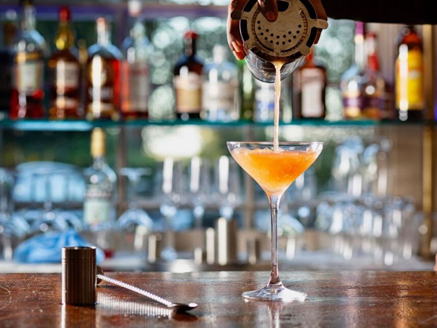 Bartender pouring a frothy cocktail into a martini glass at a polished wooden bar with spirits and glassware in the background