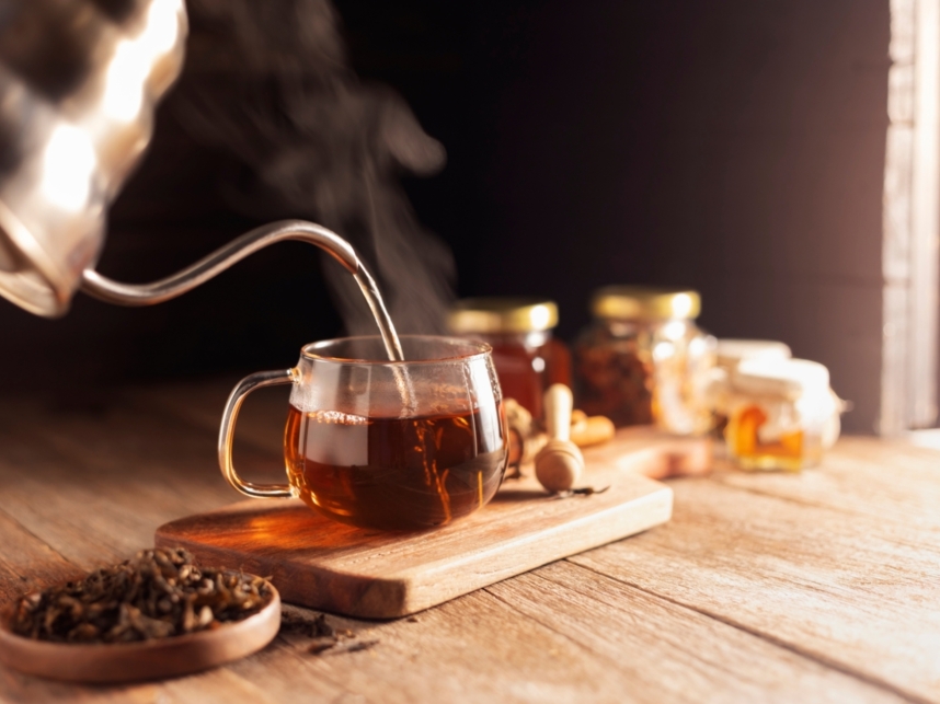 Steam rising as hot tea is poured from a kettle into a clear glass cup on a wooden board, surrounded by honey jars and loose tea leaves