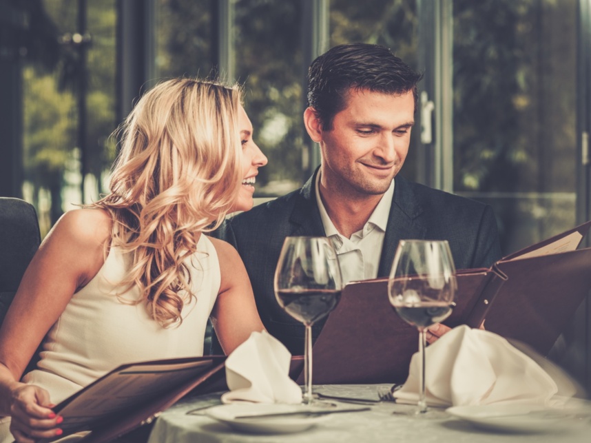 Couple dressed elegantly browsing menus at a table set with wine glasses and white napkins in a cozy restaurant