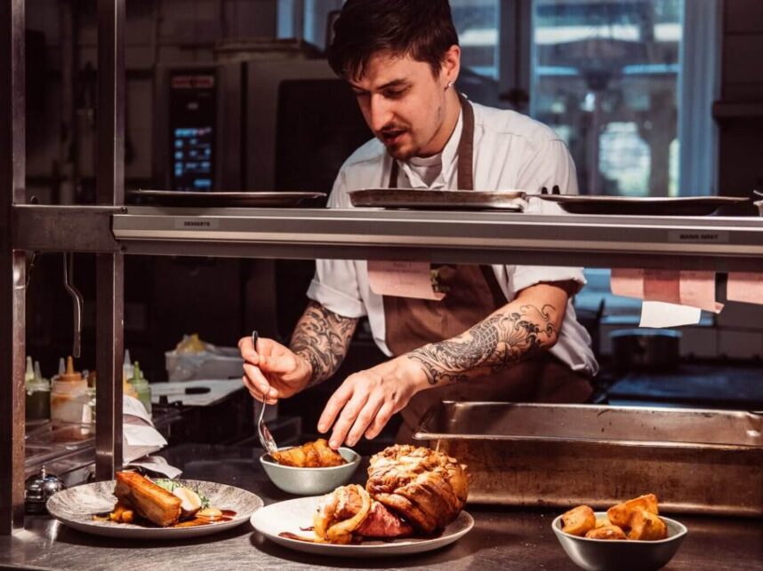 Tattooed chef plating gourmet dishes in professional kitchen, preparing a variety of meats and sides for hotel dining service