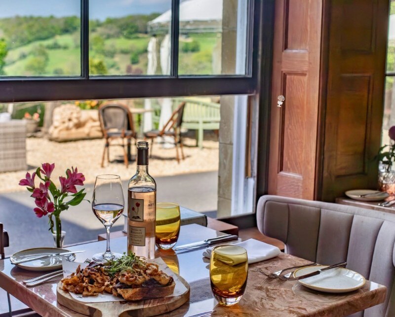 Cozy indoor dining table with wine, gourmet meal on wooden board, fresh flowers, and outdoor countryside view through large window