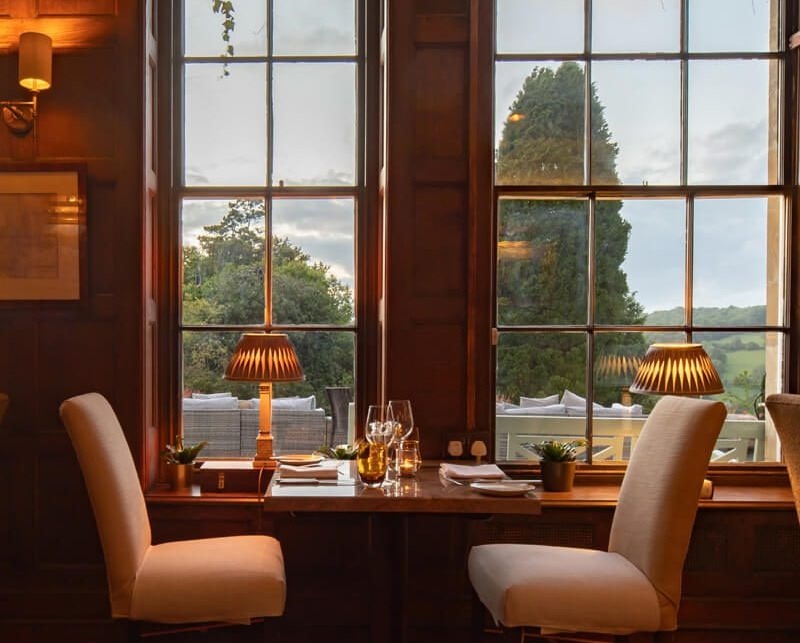 Cozy dining table for two by tall windows with soft lamps, overlooking lush greenery and outdoor seating at sunset.