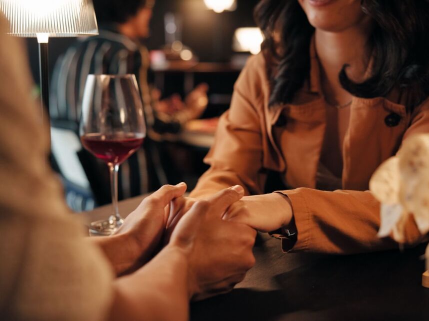 Couple holding hands across a dimly lit table with a glass of red wine, enjoying a romantic dinner in a cozy restaurant.