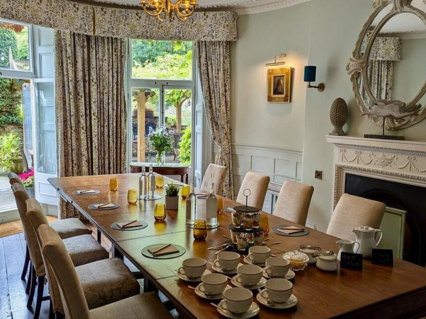 Elegant dining room with long wooden table, beige upholstered chairs, chandelier, ornate mirror, set for tea with cups and pastries, natural light