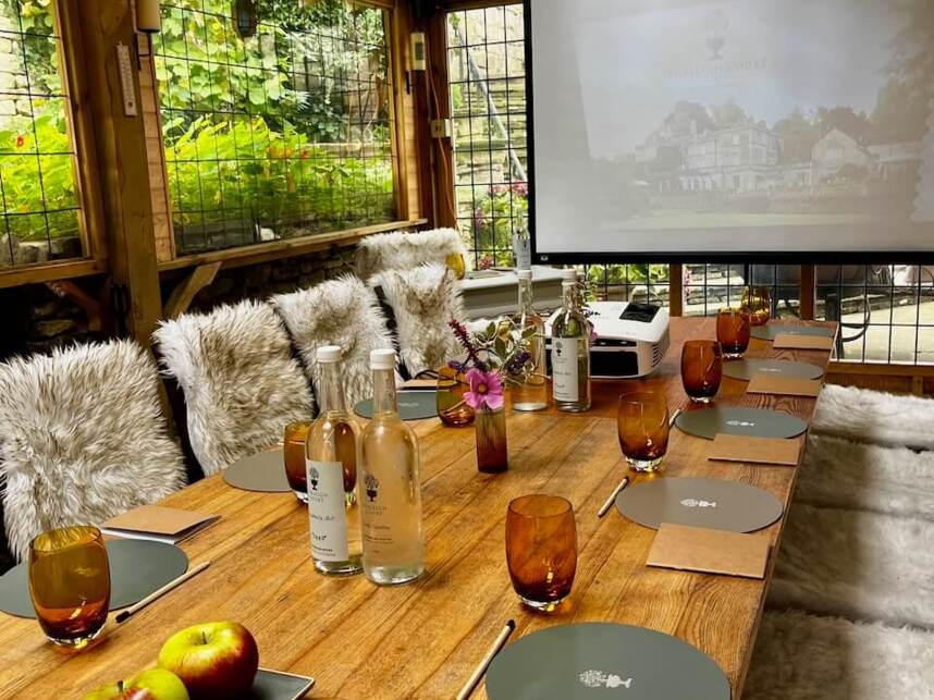 Cozy wooden meeting room with furry chair covers, amber glasses, apples, bottled water, notepads, and a projector screen in garden setting