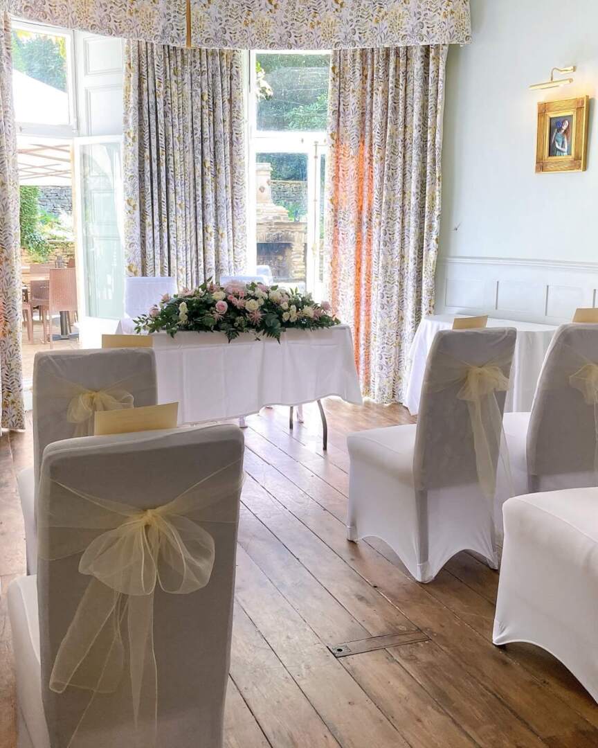 Bright wedding ceremony room with white chairs draped in sheer bows, floral centerpiece, wooden floors, and floral curtains overlooking garden.