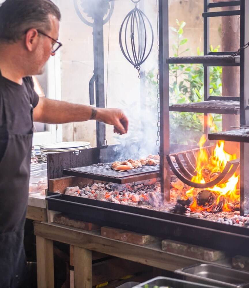 Chef grilling skewers over an open flame grill with smoke rising in a rustic outdoor kitchen setting