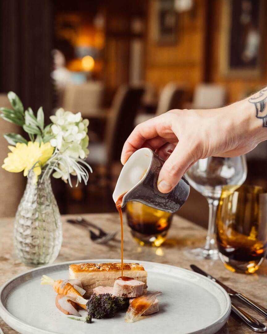 Hand pouring sauce over a plated gourmet pork dish with roasted vegetables in an elegant dining setting with glassware and fresh flowers