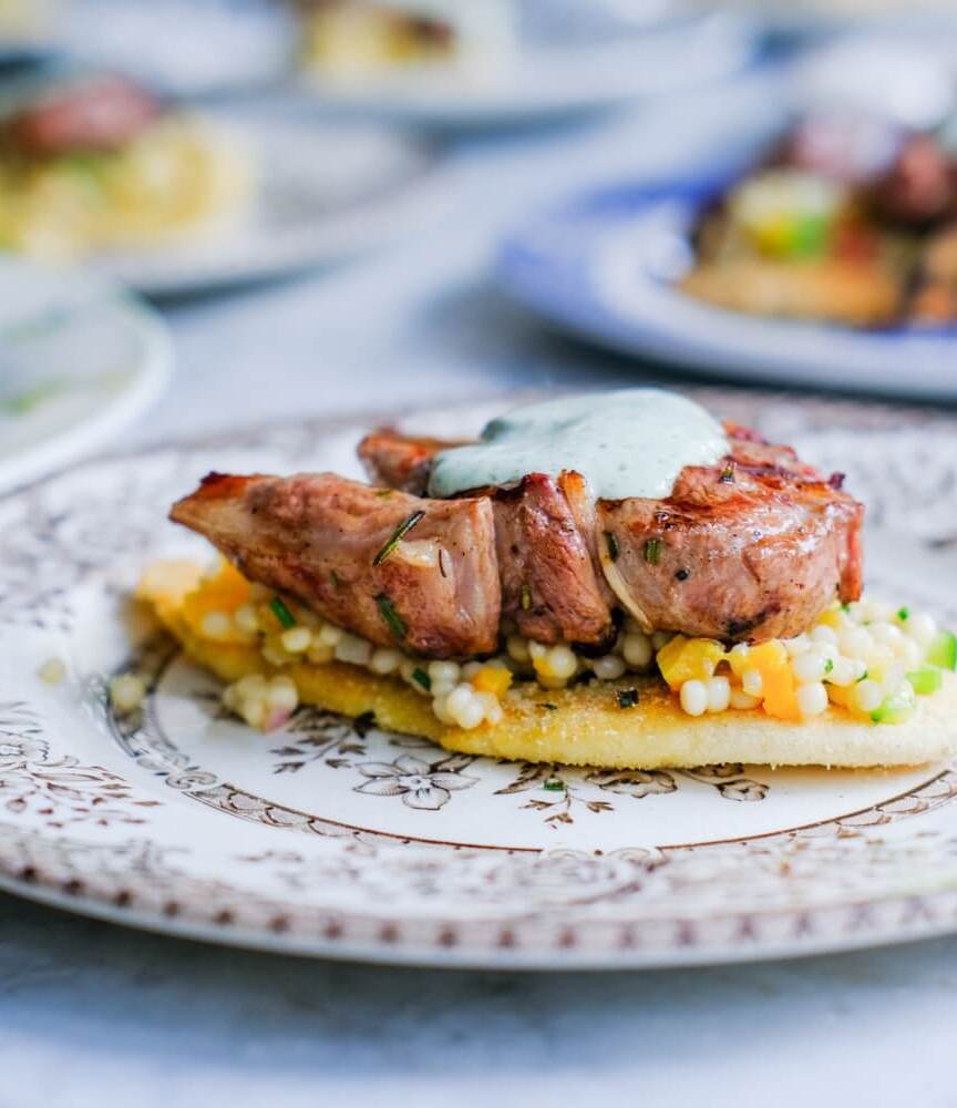 Grilled meat topped with herb sauce served on a bed of pearl couscous and vegetables on a decorative plate