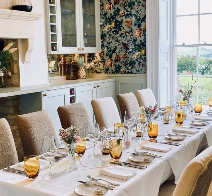 Elegant dining room with long table set for guests, amber glassware, floral centerpieces, cozy chairs, and large window overlooking green landscape