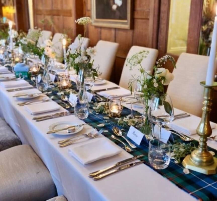 Elegant dining table set with white linens, glassware, fresh floral arrangements, and brass candlesticks in a wood-paneled room