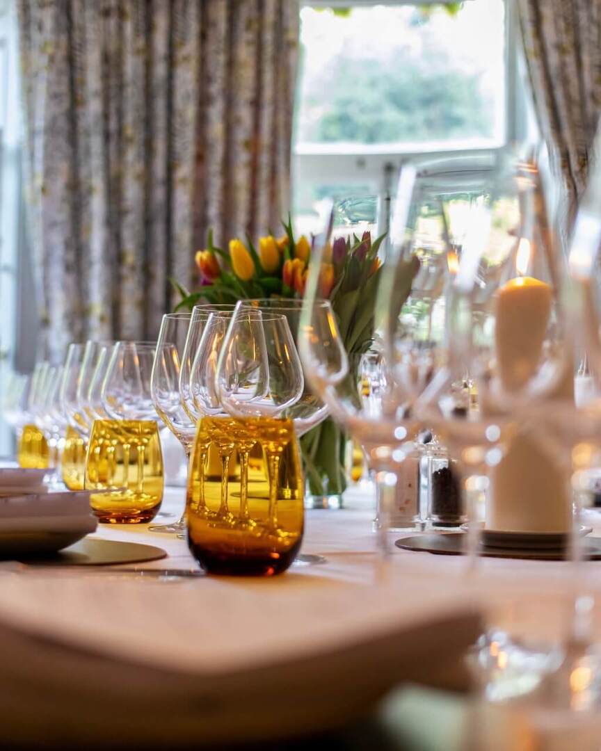 Elegant dining table set with folded napkins, amber glassware, wine glasses, candles, and a floral centerpiece near large windows