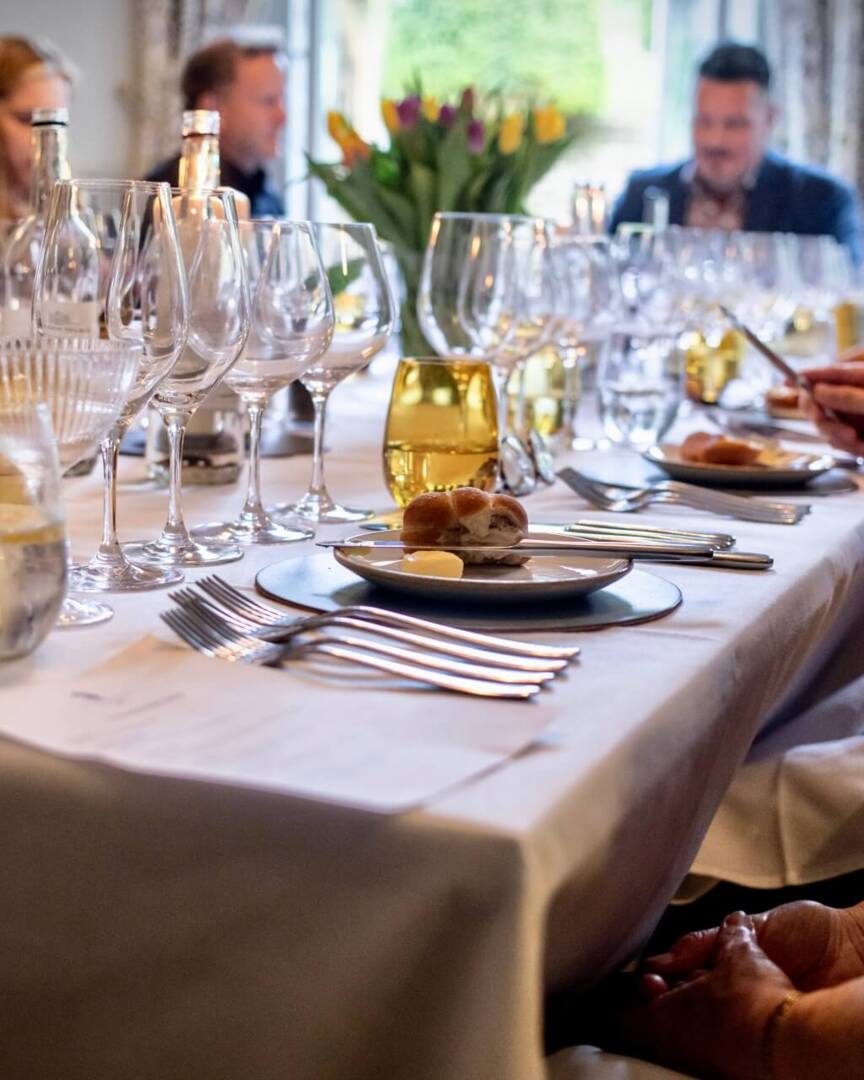 Elegant dining table set with multiple wine glasses, bread rolls, and butter, guests engaged in conversation in a cozy restaurant.