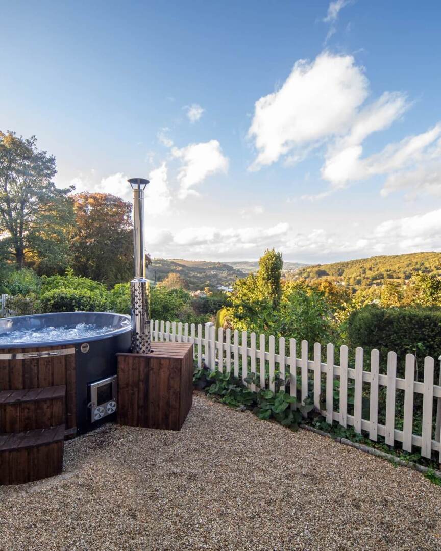Outdoor bubbling hot tub with wooden steps beside a white picket fence overlooking lush green hills under a blue sky