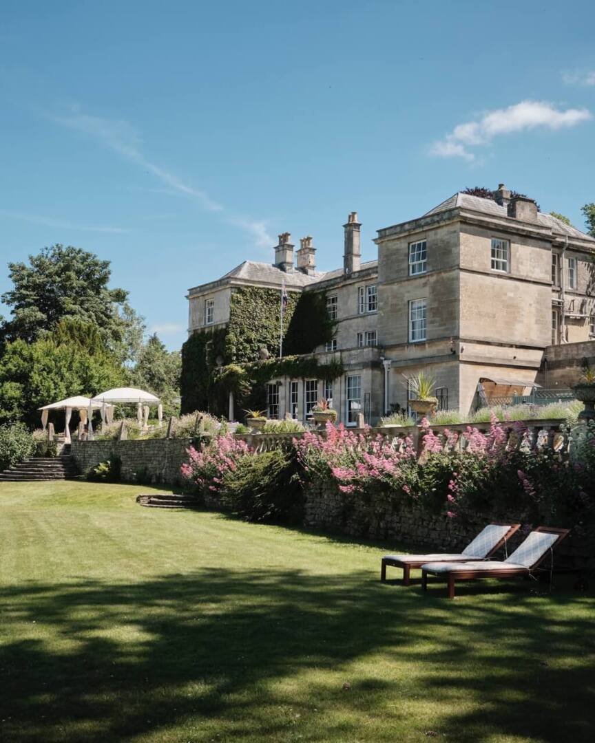 Historic stone hotel surrounded by lush gardens, pink flowers, and sun loungers on a manicured lawn under a clear blue sky