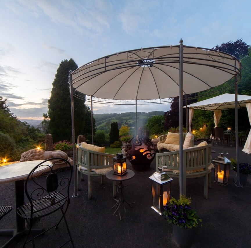 Outdoor seating area with cushioned benches under canopy, lanterns, fire pit, and scenic garden views at dusk