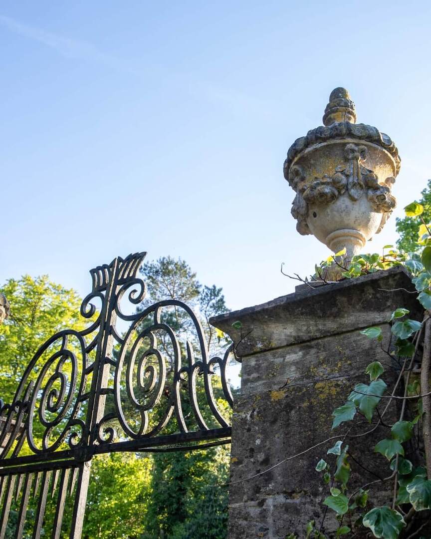 Entrée historique de l'hôtel avec portail en fer forgé décoratif, piliers en pierre surmontés d'urnes et jardin recouvert de lierre