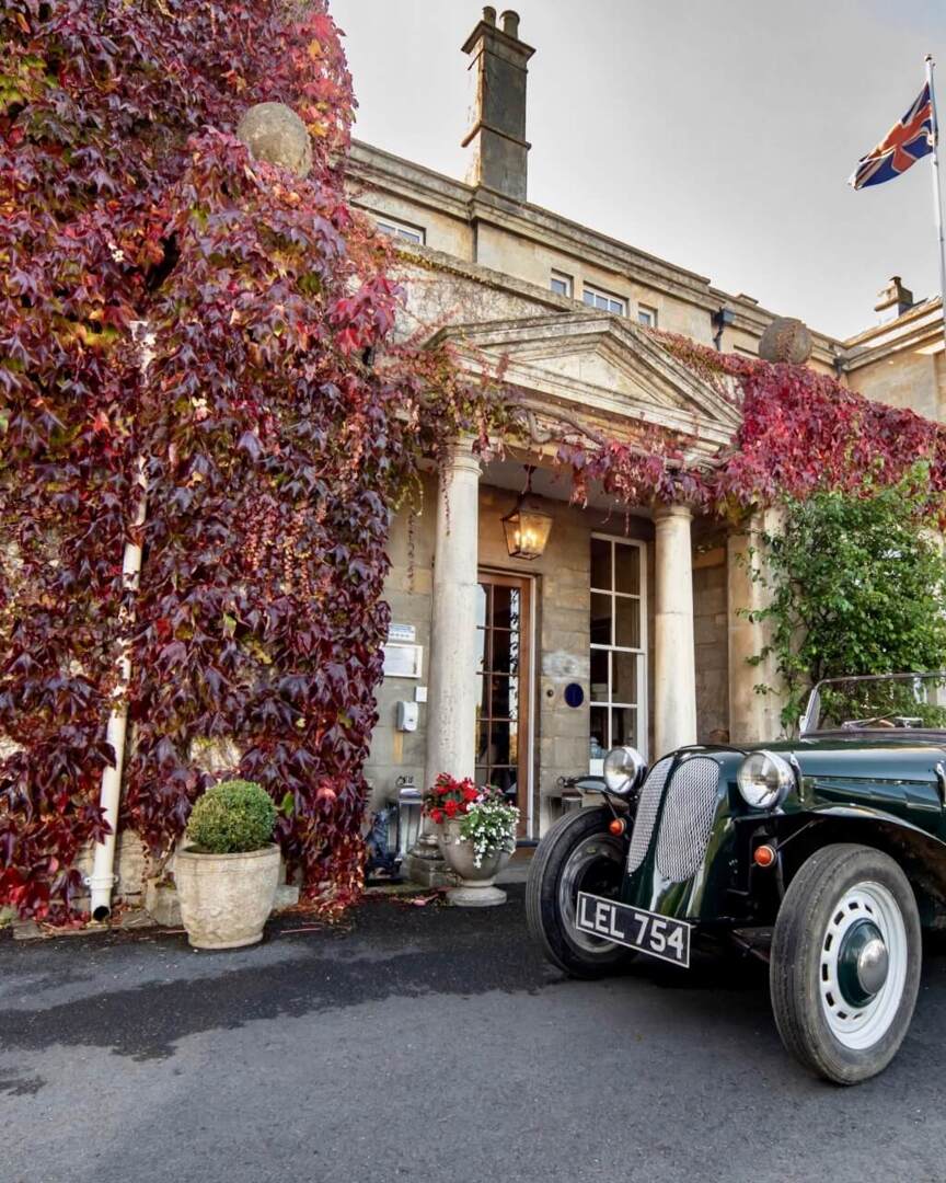 Historic stone hotel entrance with red ivy, classic convertible car parked outside, and British flag fluttering above the building