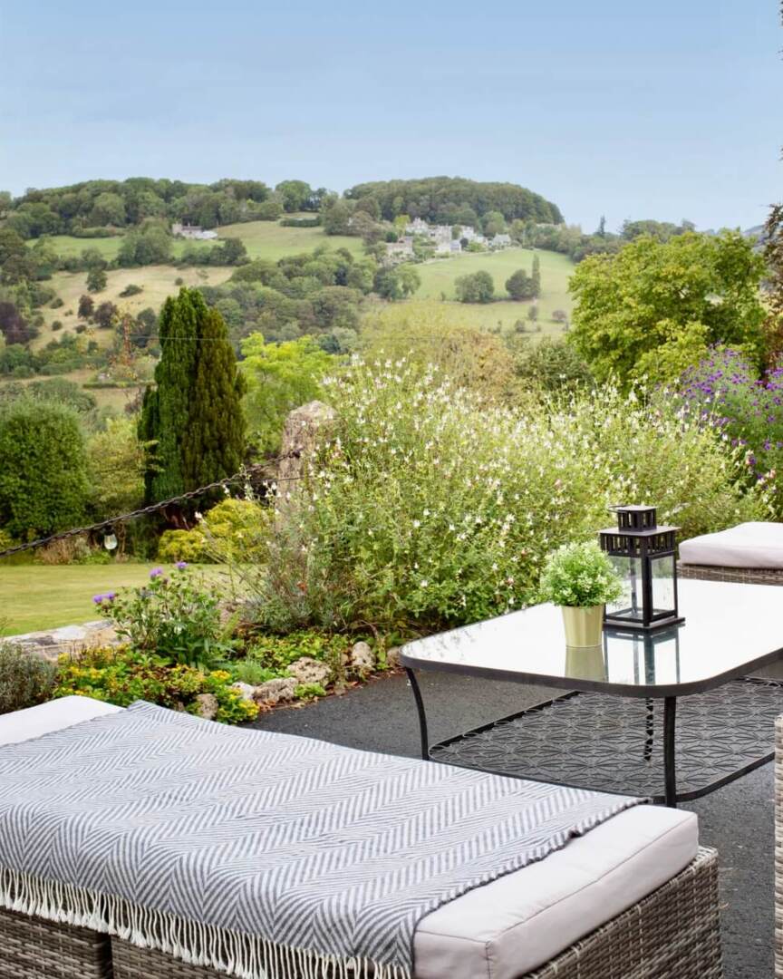 Outdoor patio with cushioned wicker seating, glass coffee table, decorative lantern, and scenic hillside views surrounded by lush greenery