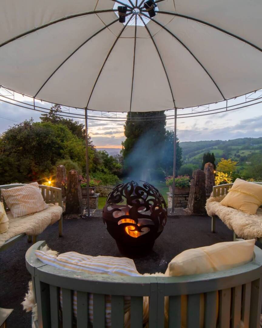 Outdoor lounge area with cushioned benches, decorative fire pit, lanterns, and canopy overlooking a scenic garden and hillside at sunset