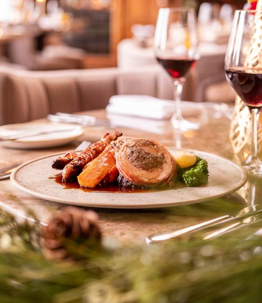 Gefüllte Bratenrolle mit glasierten Karotten und Gemüse auf elegantem Tisch, Glas Rotwein und gemütliches Restaurantambiente