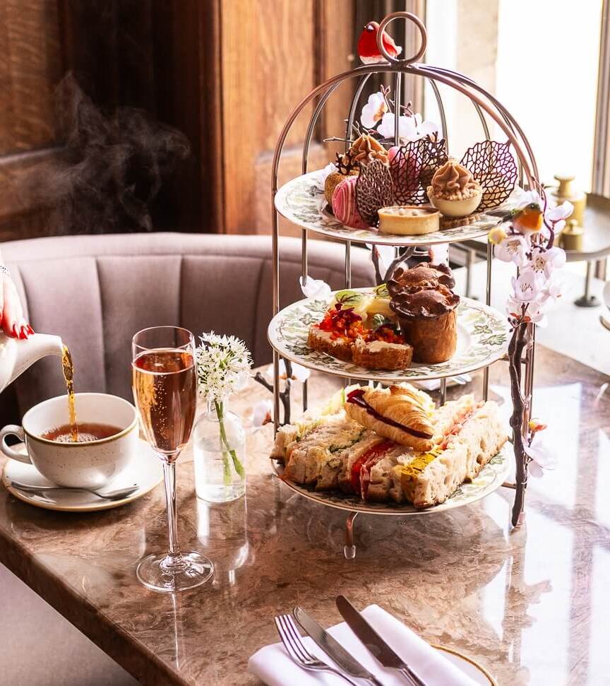 Afternoon tea setup with tiered platter of sandwiches, pastries, and desserts, steaming tea poured into a cup, and two glasses of sparkling wine