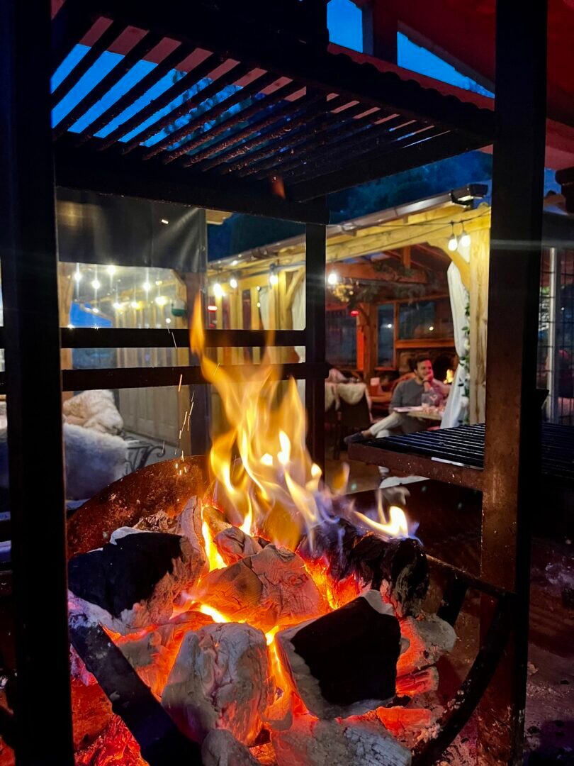 Cozy outdoor fire pit with glowing embers in a wooden pergola area, guests dining and relaxing at night nearby