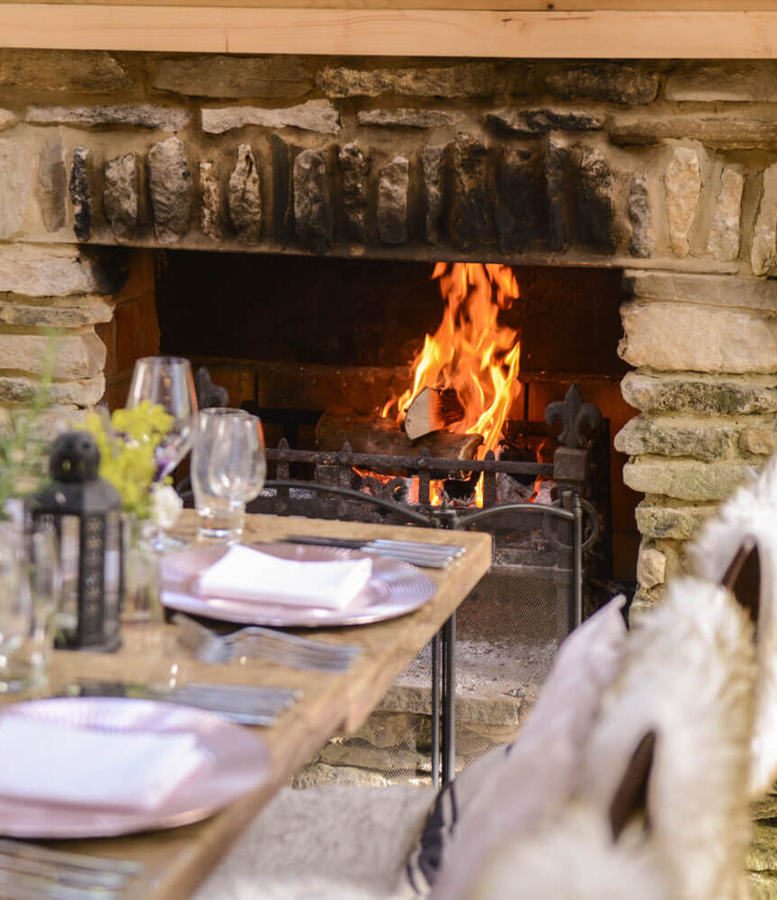 Cozy dining table set with glassware and plates by a rustic stone fireplace with a warm fire and wood stack nearby