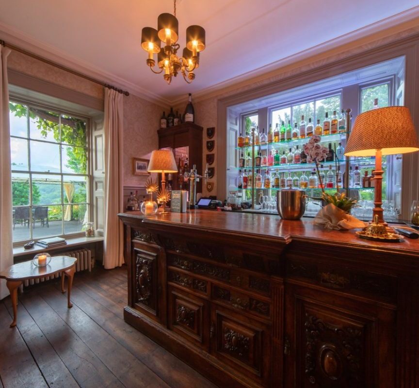 Cozy hotel bar with antique wooden counter, warm lighting, stocked liquor shelves, and a large window overlooking a green terrace