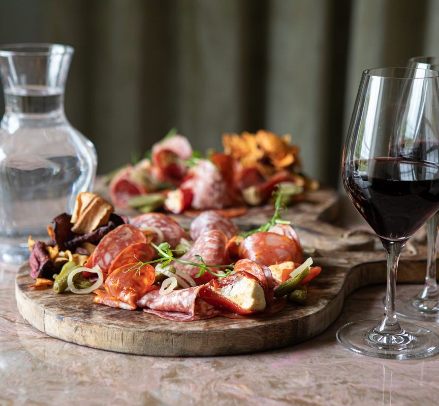 Charcuterie board with assorted cured meats, pickles, and garnishes paired with two glasses of red wine and a carafe of water on a marble table