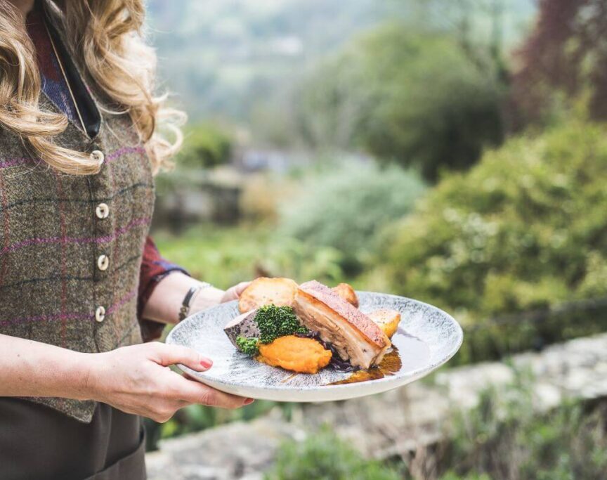 Server holding a plate with roasted pork, mashed sweet potatoes, broccoli, and roasted potatoes in a garden setting