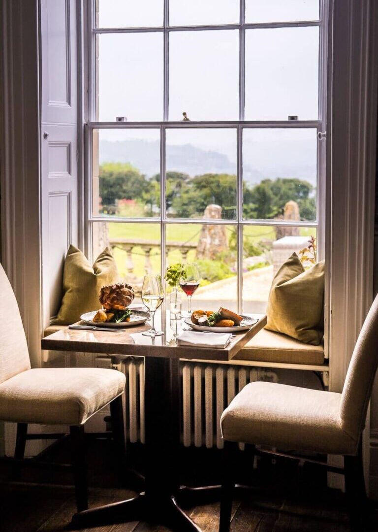 Cozy dining nook with cushioned window seats, two chairs, plates of gourmet food, wine glasses, overlooking lush garden and countryside.