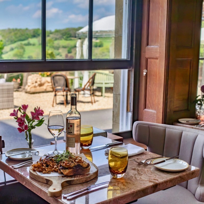 Cozy indoor dining table with wine, gourmet meal on wooden board, fresh flowers, and outdoor countryside view through large window