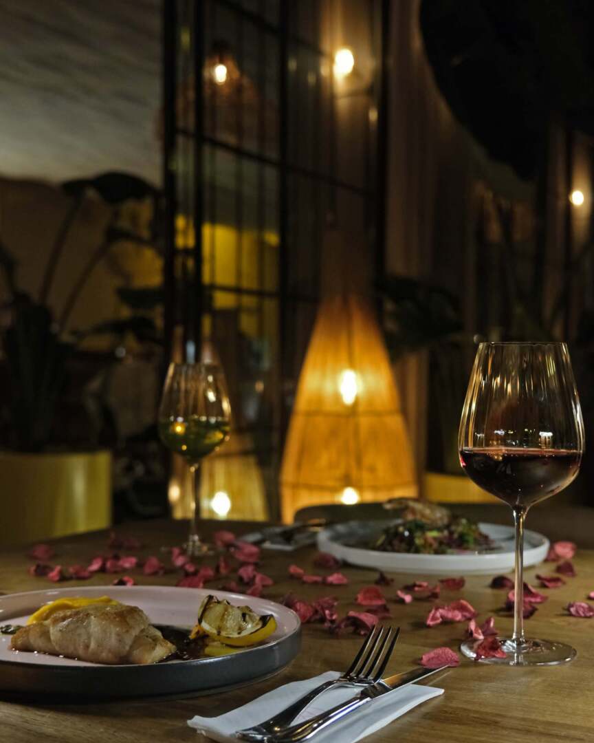 Romantic dimly lit restaurant table with gourmet dishes, red and white wine glasses, and scattered rose petals.