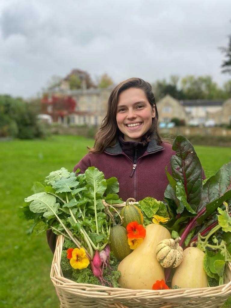 Smiling woman holds basket of fresh garden vegetables and vibrant flowers in outdoor hotel garden area