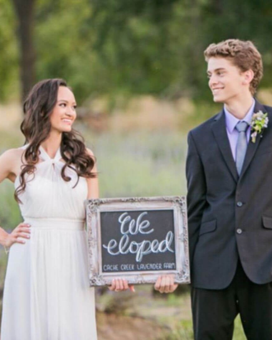 Bride in white dress and groom in suit holding a framed sign reading 'We eloped' in a scenic outdoor lavender field