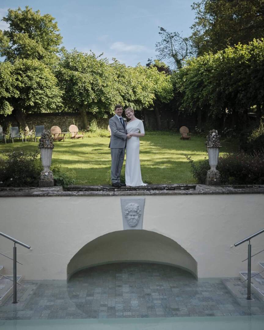 Couple in wedding attire standing by a garden terrace overlooking a decorative pool with mosaic tiles under soft daylight