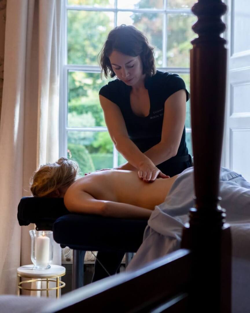 Relaxing spa massage in a softly lit room with floral wallpaper, large window, fresh flowers, and a lit candle nearby